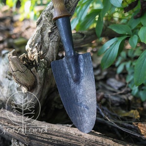 May include: A black metal garden trowel with a wooden handle. The trowel is resting on a piece of weathered wood.