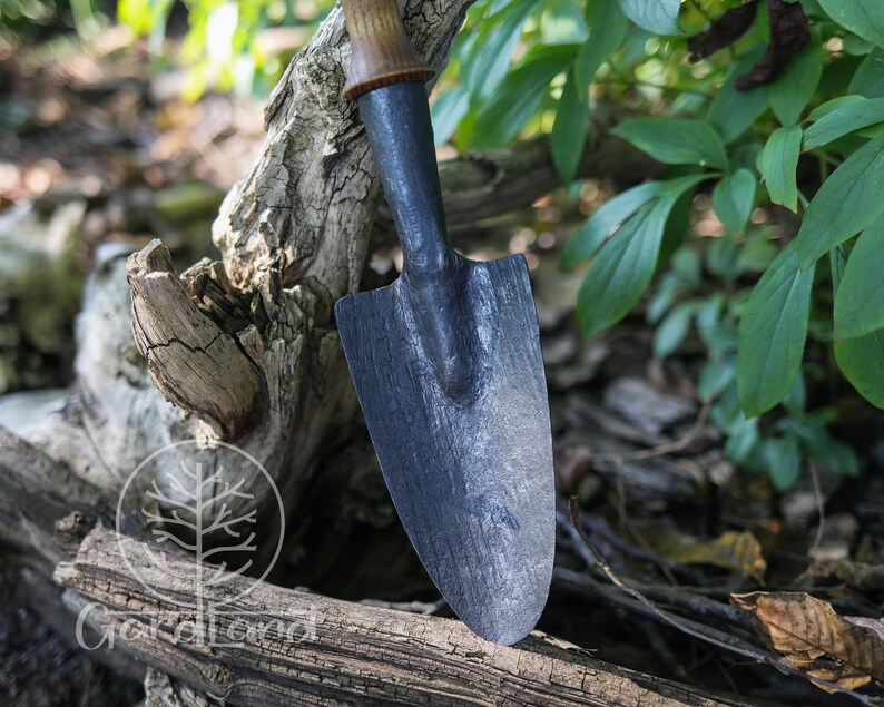May include: A black metal garden trowel with a wooden handle. The trowel is resting on a piece of weathered wood.
