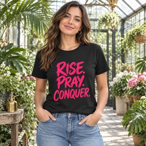 Puede incluir: Una camiseta negra de manga corta con el texto "RISE. PRAY. CONQUER." impreso en una fuente de trazo de pincel rosa brillante en el pecho, combinada con unos vaqueros azules clásicos con un botón metálico y detalles de bolsillos visibles.