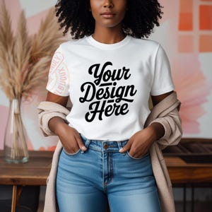 May include: A white t-shirt with the text "Your Design Here" in black lettering. The shirt is being worn by a person with dark curly hair and is styled with a light brown cardigan and blue jeans.