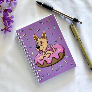 Pode incluir: Caderno roxo de espiral com um cão de desenho animado usando um donut rosa como chapéu. O cão está sentado em um donut rosa com granulado em um fundo roxo.