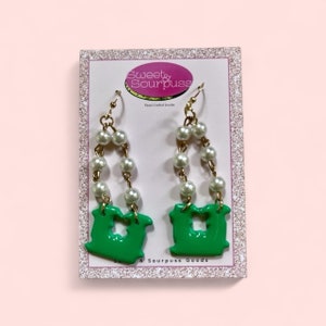 May include: Green letter U earrings with a pearl and gold chain. The earrings are on a white card with the text "Sweet & Sourpuss Hand Crafted Jewelry & Sourpuss Goods".