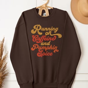Puede incluir: Una sudadera marrón con el texto "Running on Caffeine and Pumpkin Spice" en letras retro naranjas y doradas. La sudadera está colgada de una percha de madera. Un sombrero color canela es visible en el fondo.