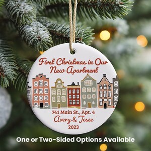 May include: A white ceramic Christmas ornament with a festive design. The ornament features a row of colorful houses and the text "First Christmas in Our New Apartment." It includes the address, names, and the year 2023. A jute string is attached for hanging.