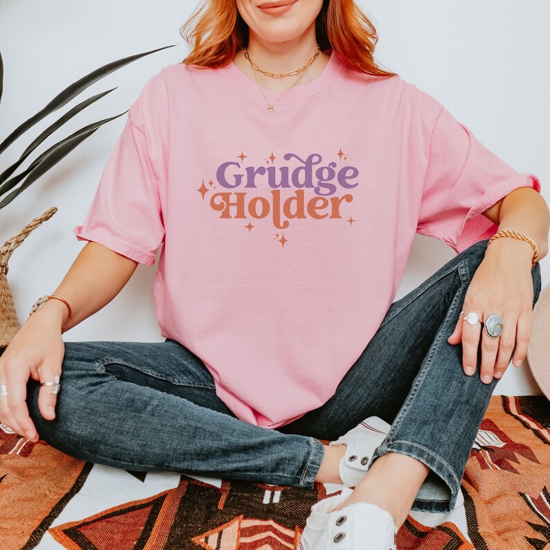 May include: A pink t-shirt with the text "Grudge Holder" in purple and orange letters with sparkly accents.