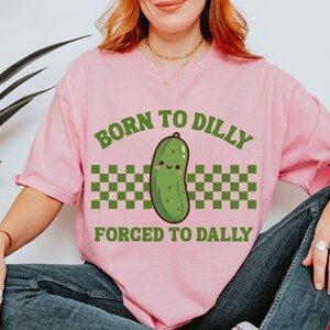 May include: A pink t-shirt featuring the text "BORN TO DILLY FORCED TO DALLY" in green. The design includes a cartoon pickle with a smiling face, above a green and white chequered pattern.