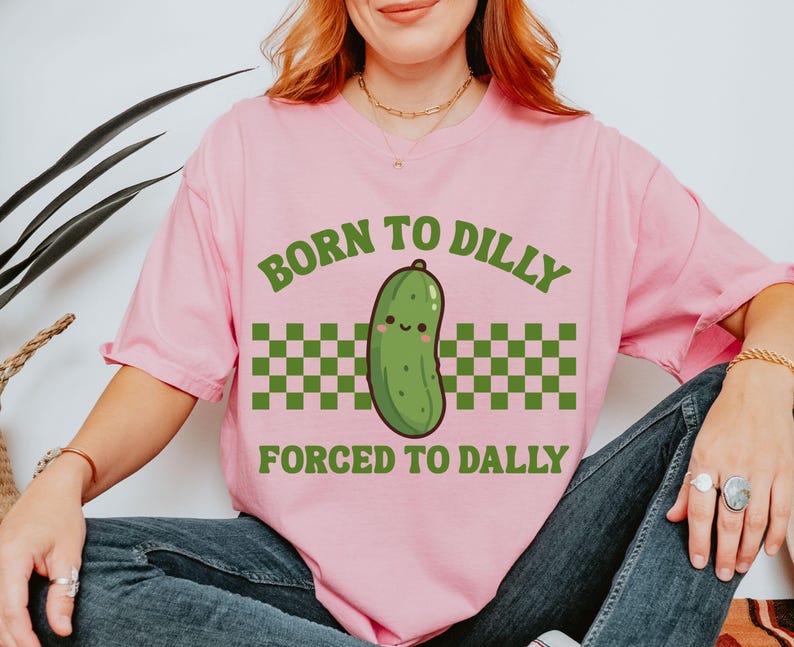 May include: A pink t-shirt featuring the text "BORN TO DILLY FORCED TO DALLY" in green. The design includes a cartoon pickle with a smiling face, above a green and white chequered pattern.