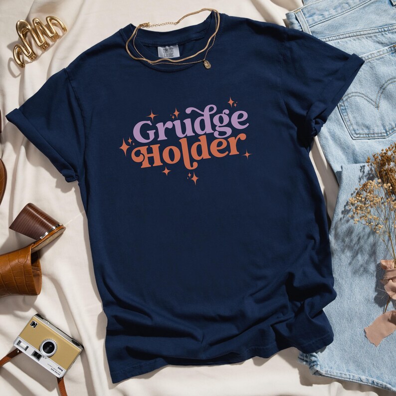 May include: A navy blue t-shirt with the text "Grudge Holder" in a retro font with pink and orange accents and sparkling stars.
