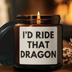 Può includere: Una candela accesa in un barattolo di vetro marrone con coperchio nero. L'etichetta sul barattolo recita "I'D RIDE THAT DRAGON" in grassetto nero. La candela è posta su una superficie di legno.