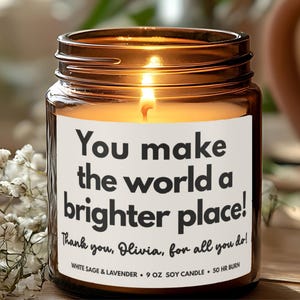 May include: A lit brown glass jar candle with a white label. The label reads "You make the world a brighter place!" with additional text. The candle is 9 oz and has a 50-hour burn time. White flowers are in the background.