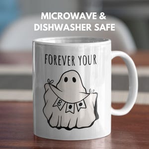 May include: White ceramic mug with a black ghost illustration holding a banner that says "BOO". The mug has the text "FOREVER YOUR" above the ghost and "MICROWAVE & DISHWASHER SAFE" above the mug. The mug has a white handle.