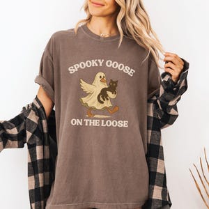 May include: Brown t-shirt with the text "Spooky Goose on the Loose." The graphic features a ghost-like goose holding a black cat. The shirt has rolled-up sleeves and is worn with a black and tan plaid shirt.
