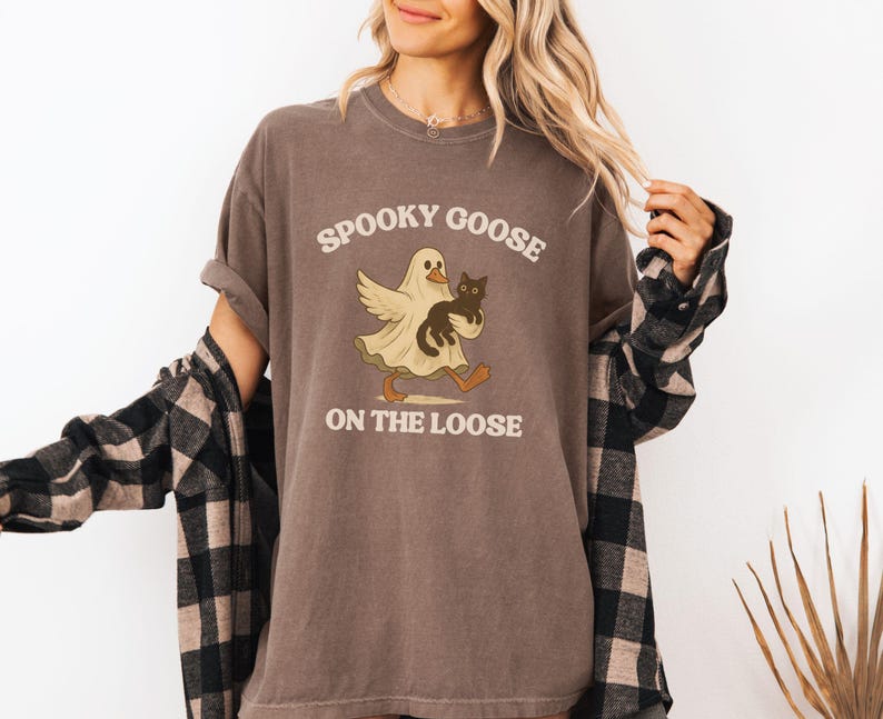 May include: Brown t-shirt with the text "Spooky Goose on the Loose." The graphic features a ghost-like goose holding a black cat. The shirt has rolled-up sleeves and is worn with a black and tan plaid shirt.