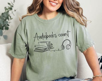 May include: Sage green t-shirt featuring the phrase "Audiobooks count" in a handwritten style. Below the text is a line drawing of an open book and headphones. The t-shirt has a relaxed fit.