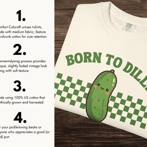 May include: Cream-coloured unisex t-shirt with the phrase "BORN TO DILLY" in green lettering, featuring a smiling cartoon pickle. The shirt has a green and white checkered pattern and is made of medium fabric.