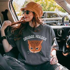 May include: A gray t-shirt with the text "GRUDGE HOLDER" and a cartoon image of an orange cat with green eyes.