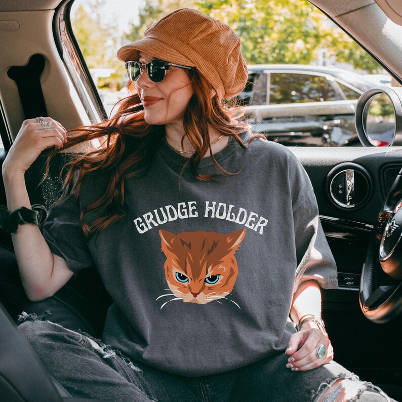 May include: A gray t-shirt with the text "GRUDGE HOLDER" and a cartoon image of an orange cat with green eyes.