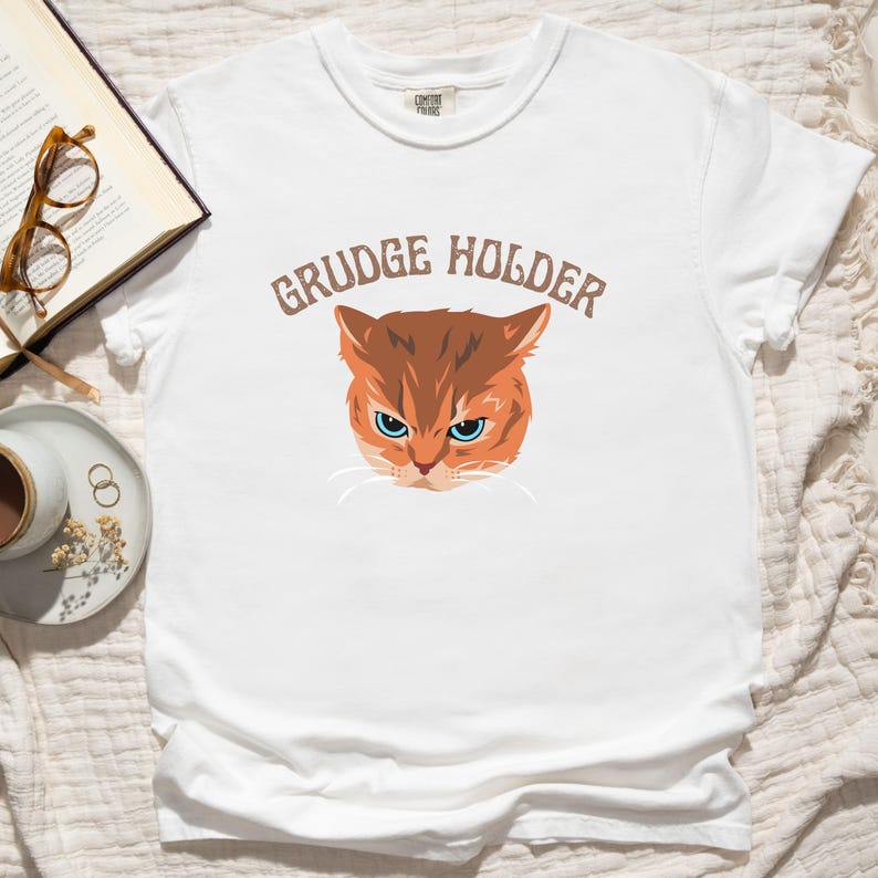 May include: White t-shirt with a brown cat face and the text "GRUDGE HOLDER" in brown letters.