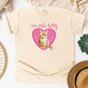 Op de afbeelding: Een beige T-shirt met een roze hartvormig ontwerp met een schattig oranje kitten. De tekst "one cute kitty!" staat boven het hart.