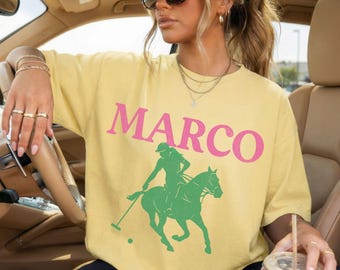 Könnte beinhalten: Hellgelbes T-Shirt mit dem Wort "MARCO" in rosa Blockbuchstaben. Darunter befindet sich eine grüne Silhouette eines Polospielers auf einem Pferd. Das Shirt hat eine lockere, übergroße Passform.