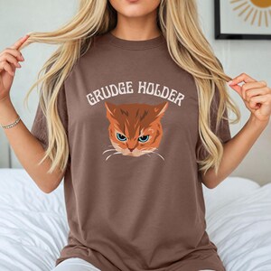 May include: Brown t-shirt with a graphic of an orange cat with the text "GRUDGE HOLDER" printed in white.