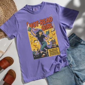 Pode incluir: Uma camiseta lilás com um gráfico em estilo de banda desenhada. O gráfico diz "HOT-READ GIRL" e apresenta uma mulher segurando livros. O gráfico também inclui o texto "ARE THESE OUR DAUGHTERS?" e outro texto.