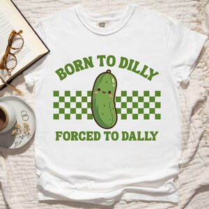 May include: White t-shirt with the text "BORN TO DILLY FORCED TO DALLY" in green. The shirt features a cartoon pickle illustration with a smiling face, set against a green and white checkered pattern.