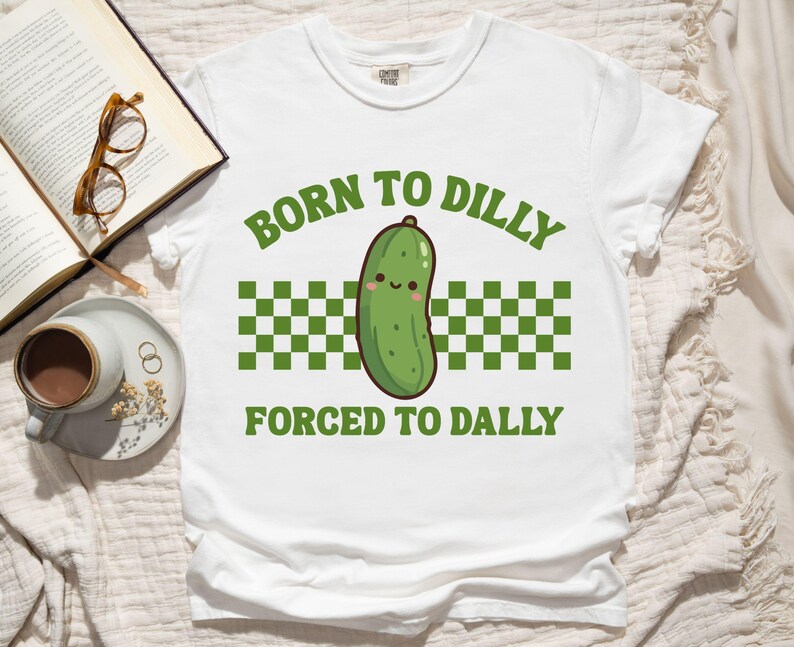 May include: White t-shirt with the text "BORN TO DILLY FORCED TO DALLY" in green. The shirt features a cartoon pickle illustration with a smiling face, set against a green and white checkered pattern.