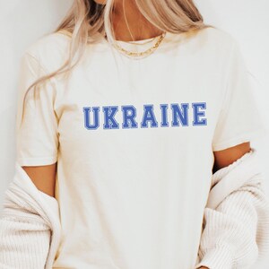 以下が含まれることがあります： 白いTシャツに、大学のフォントで書かれた青い「UKRAINE」のグラフィックがプリントされています