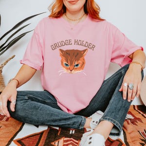 May include: A pink t-shirt with a cartoon cat and the text "GRUDGE HOLDER" printed on the front.