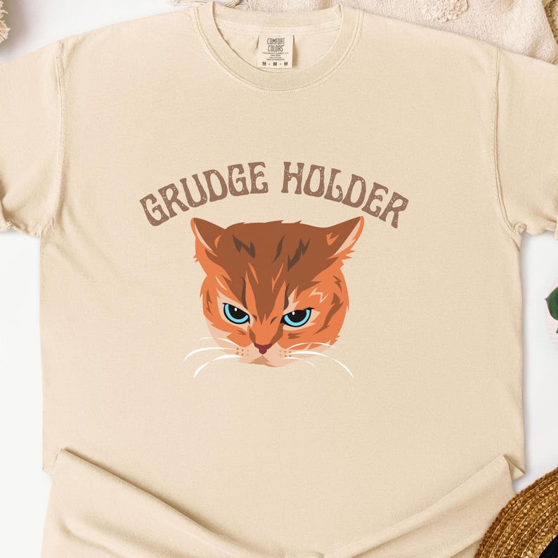 May include: A beige t-shirt with the text "GRUDGE HOLDER" in brown lettering. A cartoon orange cat with blue eyes is featured below the text.