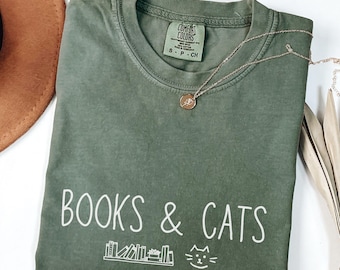 Może przedstawiać: Oliwkowo-zielona koszulka z białym napisem „BOOKS & CATS”. Poniżej tekstu znajduje się grafika książek i kota. Złoty naszyjnik z wisiorkiem spoczywa na koszulce. Koszulka jest złożona na białej powierzchni.
