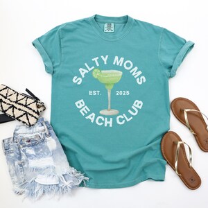 Op de afbeelding: Turquoisekleurig T-shirt met de tekst "SALTY MOMS BEACH CLUB" rond een margarita-afbeelding. Het ontwerp bevat "EST. 2025". Het shirt wordt gecombineerd met een spijkerbroekshort, een zwart-witte clutch en bruine sandalen met gouden bandjes. Thema: strandkleding.