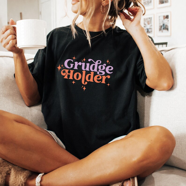 May include: A black t-shirt with the text "Grudge Holder" in a retro font with pink and orange accents and small white stars.