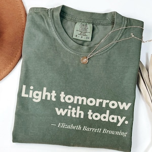 May include: An olive green t-shirt with the quote "Light tomorrow with today." by Elizabeth Barrett Browning in white lettering. A gold necklace and a brown hat are also visible.
