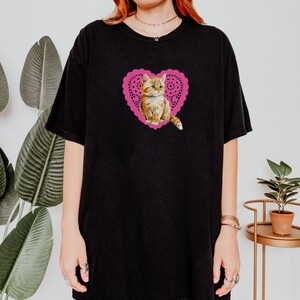 May include: Black oversized t-shirt with a pink heart-shaped doily featuring a cute orange tabby kitten.