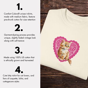 May include: A white t-shirt with a pink heart-shaped design featuring a cartoon kitten. The text "Comfort Colors&reg; unisex t-shirts, made with medium fabric, feature pre-shrunk cotton for size retention" is on the left side of the image. The text "Garment-dyeing process provides unique, slightly faded vintage look along with soft texture" is also on the left side of the image. The text "Made using 100% US cotton that is ethically grown and harvested" is on the left side of the image. The text "Cute kitty t-shirt for cat lovers, and fans of coquette, lolita, and cottagecore styles" is on the left side of the image.