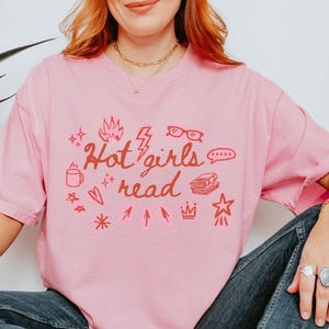 May include: A light pink t-shirt with the phrase "Hot girls read" in red script and various red illustrations. The illustrations include a coffee mug, glasses, a book, and a star.