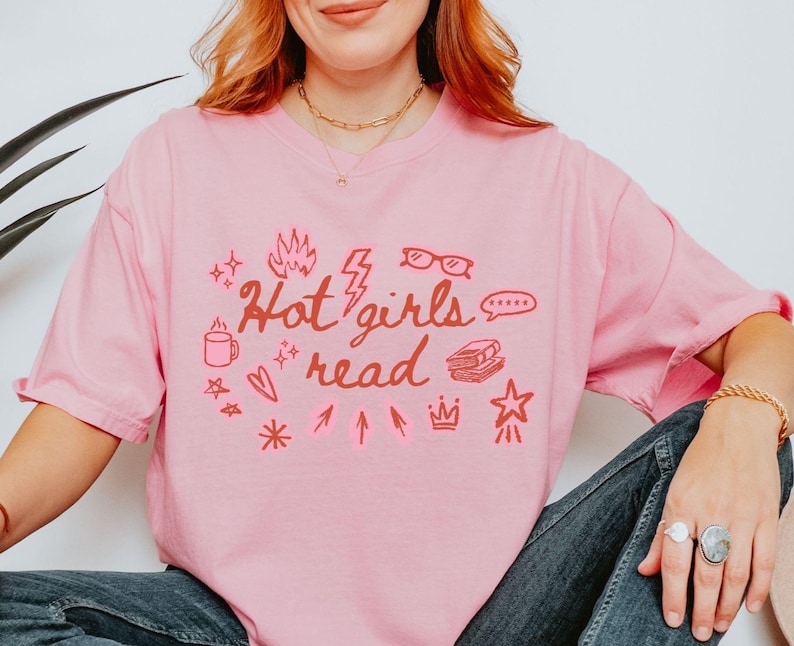 May include: A light pink t-shirt with the phrase "Hot girls read" in red script and various red illustrations. The illustrations include a coffee mug, glasses, a book, and a star.