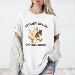 May include: White t-shirt with a graphic of a cartoon goose in a ghost costume holding a black cat. The text "Spooky Goose On The Loose" is printed above and below the graphic in a dark grey color. The shirt has short sleeves and a relaxed fit.
