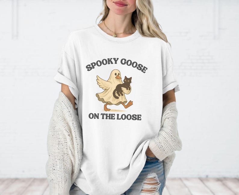 May include: White t-shirt with a graphic of a cartoon goose in a ghost costume holding a black cat. The text "Spooky Goose On The Loose" is printed above and below the graphic in a dark grey color. The shirt has short sleeves and a relaxed fit.