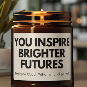 May include: A lit brown glass jar candle with a white label. The label reads "YOU INSPIRE BRIGHTER FUTURES" in bold black letters. The text "Thank you, Coach Williams, for all you do!" is below. The candle is on a wooden surface.