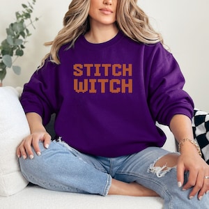May include: A purple sweatshirt with the text "STITCH WITCH" in orange embroidered letters. The sweatshirt is worn by a person sitting on a white couch.