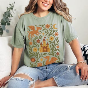 Op de afbeelding: Saliegroen T-shirt met een fantasie-ontwerp. De afbeelding bevat een kasteel, draken, bloemen en een maan in oranje, geel en bruin. Het shirt heeft korte mouwen en een relaxte pasvorm, geschikt voor dagelijks gebruik.