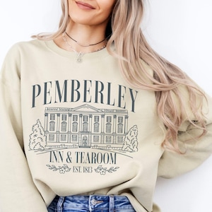 Può includere: Una felpa beige con la scritta "PEMBERLEY INN & TEAROOM EST. 1813" in blu scuro. Il design include un'illustrazione dettagliata di un edificio e dettagli floreali. La felpa è uno stile casual.