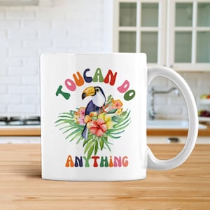 May include: White ceramic coffee mug featuring a toucan and tropical flowers. The mug displays the text "You Can Do Anything" in colourful letters.