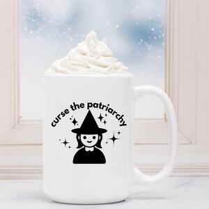 May include: White ceramic mug with a black graphic of a witch with a pointed hat and the text "curse the patriarchy". The mug has a black handle and is filled with white whipped cream.