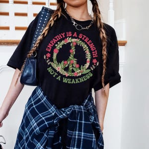 Könnte beinhalten: Schwarzes T-Shirt mit einem Peace-Zeichen-Motiv und dem Text "Empathie ist eine Stärke, keine Schwäche". Das Peace-Zeichen ist von Blumen und Blättern umgeben.