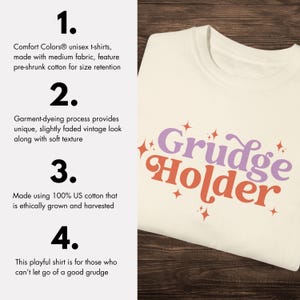 May include: A white t-shirt with the text "Grudge Holder" in pink and orange letters with a retro font. The text is surrounded by small stars. The shirt is made of 100% US cotton and is ethically grown and harvested.
