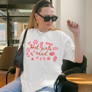 May include: White t-shirt with the text "Hot girls read" in pink, along with various pink graphic elements. The shirt is worn by a person wearing black sunglasses and a black headband.